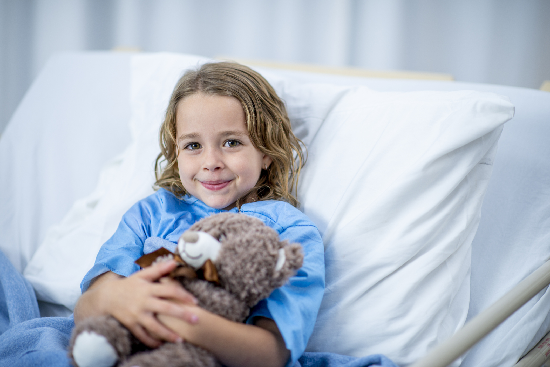 A Caucasian girl is indoors in a hospital room. She is smiling at the camera while holding a teddy bear.